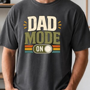 Peut inclure: T-shirt gris foncé avec l'inscription "DAD MODE ON" dans un style rétro. Le texte est en blanc cassé et vert olive, avec des accents orange et jaune. Le t-shirt a un aspect vintage et usé.