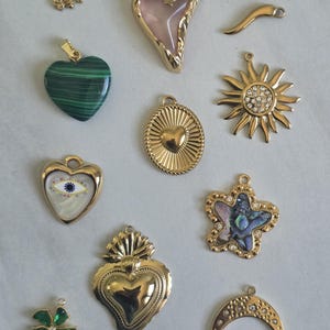 Puede incluir: Una colección de diez dijes de joyería de metal dorado que incluye una abeja, un corazón rosa, un cuerno curvo, un corazón de malaquita verde, un sol con cristales, un corazón con un ojo, un óvalo texturizado, una estrella con nácar y un corazón.