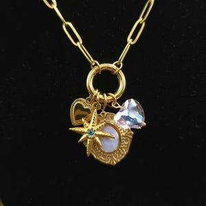 May include: Gold-coloured necklace featuring multiple charms. Charms include a heart, a starburst with a blue gem, a heart-shaped crystal, and a framed oval. The necklace has a large clasp and a chain with elongated links.