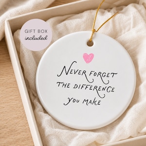 以下が含まれることがあります： 小さなピンクのハートと黒い文字で「Never forget the difference you make」と書かれた白い円形のセラミック製オーナメント。金色の吊り下げ用紐が付いており、「GIFT BOX included」というラベルの付いた箱に入っています。