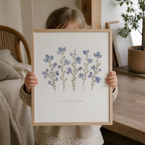 Peut inclure: Une impression encadrée avec une illustration à l'aquarelle de tiges florales bleues. L'œuvre d'art a un cadre en bois clair et le texte "Home is where mum is" en bas. L'impression est tenue par une personne.
