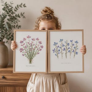 Könnte beinhalten: Zwei gerahmte Kunstdrucke mit Blumenmotiven. Ein Druck zeigt einen Strauß lila Blumen, die mit einer Schleife gebunden sind, der andere blaue Blumen. Beide sind in hellen Holzrahmen. Text ist am unteren Rand der Drucke sichtbar.