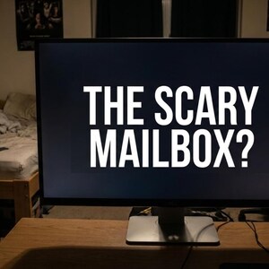 Op de afbeelding: Een grote computermonitor toont de tekst "THE SCARY MAILBOX?" in witte, vette letters tegen een donkere achtergrond. De monitor staat op een houten bureau in een kamer met een bed en posters aan de muur.