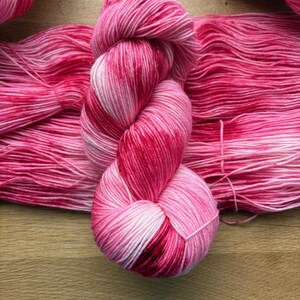 Könnte beinhalten: Ein Strang handgefärbter Wolle in Rosa- und Rottönen. Das Garn ist eng zu einem Knäuel gewickelt, mit losen Strähnen. Der Farbverlauf geht von Hellrosa zu Dunkelrot über. Ideal für Strick- und Häkelprojekte. Das Garn liegt auf einer Holzoberfläche.