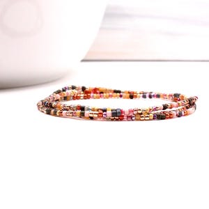 May include: A multi-strand beaded bracelet in shades of pink, gold, brown, and black. The bracelet is made of small, colourful beads strung together. The bracelet is wrapped around itself, creating a layered effect. The background is white.