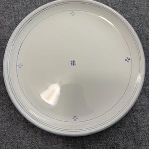 May include: A round white ceramic plate featuring a thin blue circular border near the rim. The centre of the plate is decorated with a small cluster of nine blue dots arranged in a square, with four additional dot clusters placed at intervals.