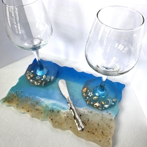 May include: Two clear wine glasses with blue stems rest on a resin tray. The tray depicts a beach scene with blue, white, and tan colours, and small seashells. A silver butter knife is also on the tray.