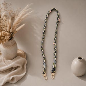 May include: A long, braided strap featuring a mix of dark gray, white, and green fabric strands interwoven with small, round, light pink beads. The strap ends in two gold-toned metal swivel lobster clasps for secure attachment to a bag or accessory.