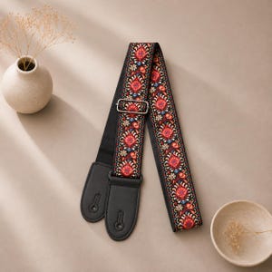 May include: A guitar strap featuring a black base with a vibrant, multi-colored floral and paisley-inspired woven pattern in shades of red, blue, and white. The strap includes black leather end tabs with keyhole slots and a silver metal slider.