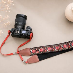 May include: A black digital camera with a lens attached to a camera strap. The strap features a vibrant floral pattern in red, pink, and white on a black background, accented with brown leather ends and thin red nylon webbing connectors.