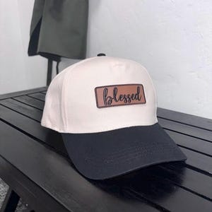 Może przedstawiać: Czapka bejsbolowa z kremową koroną i czarnym daszkiem. Czapka posiada prostokątną naszywkę z napisem "blessed" w kursywie.
