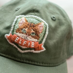 以下が含まれることがあります： 詳細な猫の顔が刺繍されたパッチが付いた、オリーブグリーンの野球帽。「ELFIE」という文字がバナーに書かれています。帽子は柔らかい生地でできており、つばがカーブしています。