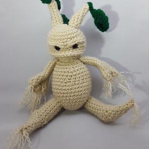 May include: A crocheted plush toy shaped like a humanoid root with green leaves. The toy has a beige body with two long arms and legs, and two green leaves on top of its head.