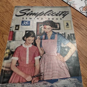 May include: Vintage "Simplicity Sewing Book" cover featuring two women, one in a red and white striped apron. The cover has the text "Simplicity Sewing Book" and "Easy Guide for Beginners and Experts". The cover is aged with a worn appearance.