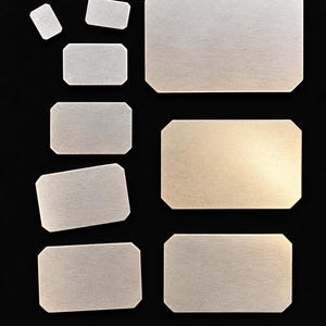 May include: A collection of eight rectangular metal plates with clipped corners, arranged in two columns. The plates feature a brushed silver finish with a subtle metallic sheen, varying in size from small to large, all displaying a flat surface.