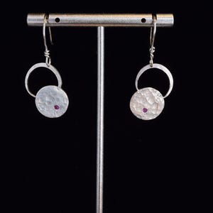 May include: Silver earrings with a hammered texture and a small, round, magenta-colored stone in the center of each. The earrings are suspended from a silver hook and a small silver ring.