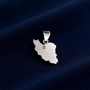 May include: A silver pendant in the shape of Iran, with a small, round, yellow gemstone set in the center. The pendant hangs from a silver bail. The background is a dark blue, textured fabric.