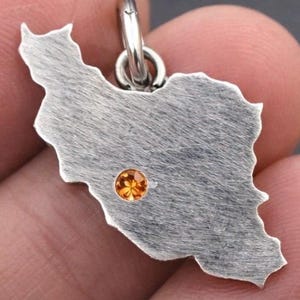 May include: A silver-colored pendant in the shape of Iran, with a small, round, orange gemstone embedded in the center. The pendant is attached to a silver loop.