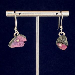 May include: A pair of dangle earrings featuring rough-cut pink tourmaline gemstones with dark gray matrix. The earrings are suspended from silver-colored hooks and displayed on a silver earring stand against a dark blue background.