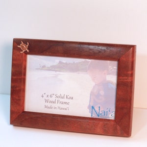 May include: A rectangular solid Koa wood frame, 4 inches by 6 inches, with a small turtle accent in the upper left corner. The frame holds a photo of a child on a beach. Text on the frame reads "4" x 6" Solid Koa Wood Frame Made in Hawai'i".