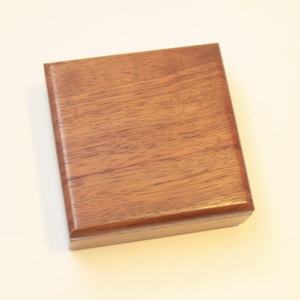 May include: A square, wooden box with a rich, brown finish. The wood grain is visible, adding texture to the surface. The box has a smooth, polished appearance, suggesting it is well-crafted. The edges are slightly rounded.
