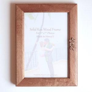 May include: A rectangular solid Koa wood frame for 5"x7" photos. The frame has a natural wood grain finish and features two small wooden turtle embellishments on the right side. Text on the frame reads "Solid Koa Wood Frame" and "Made in Hawai'i".