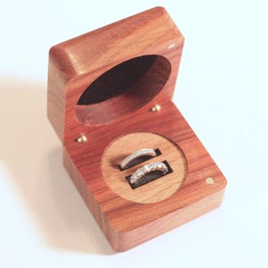 May include: Open wooden ring box with two diamond rings. The box is made of reddish-brown wood and has a circular inset with a black velvet lining. The box is open, revealing the rings.