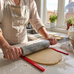 May include: A grey marble rolling pin with wooden handles rolls out dough between two red guides. The guides are marked "6mm (1/4")". Cookie cutters and baked cookies are in the background. The scene takes place in a well-lit kitchen.