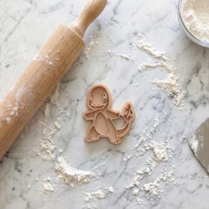 Puede incluir: Cortador de galletas Charmander de plástico marrón claro sobre una superficie de mármol espolvoreada con harina. También se ven un rodillo de madera, un cuenco de harina y una espátula de metal. El cortador de galletas tiene la forma del personaje Pokémon.