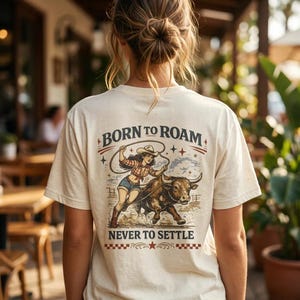 Può includere: Una t-shirt color crema con una grafica sul retro. Il disegno mostra una persona con cappello da cowboy, camicia a quadri e pantaloncini di jeans che cavalca un toro tenendo un lazo. Il testo riporta BORN TO ROAM e NEVER TO SETTLE.