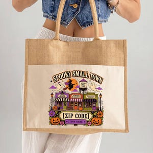 May include: A rectangular jute tote bag with a cream-colored canvas front panel featuring a colorful graphic of a spooky small town. The design includes a witch on a broom, ghosts, a water tower, and shops with the text Spooky Small Town Zip Code.