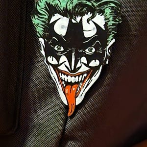 Puede incluir: Un llavero de metal con una cara estilizada con pelo verde, una máscara negra y una lengua roja. El diseño recuerda a un villano de cómic. El llavero está sujeto a un anillo plateado.