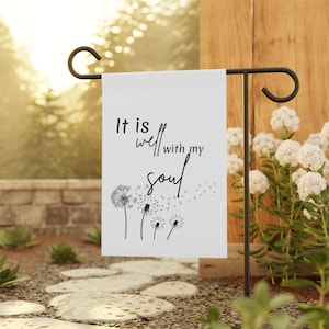 May include: A white rectangular garden flag hanging on a black metal stand. The flag features black text that reads It is well with my soul, accompanied by a black graphic illustration of four dandelion seed heads blowing in the wind.