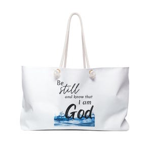 May include: White tote bag with rope handles. The bag features the text "Be still and know that I am God" in black script and a blue water graphic.