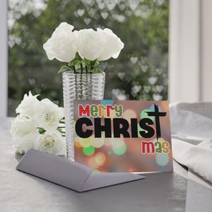 May include: A rectangular greeting card featuring the text MERRY CHRISTmas with a black cross replacing the letter T, set against a blurred bokeh background. A clear glass vase holds a bouquet of white roses next to the card.