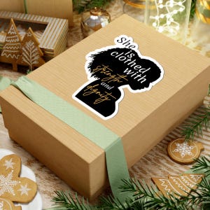 May include: A die-cut sticker featuring a black silhouette of a person's head and shoulders with the white and gold text "She is clothed with strength and dignity" placed on a light brown rectangular gift box wrapped with a green ribbon.