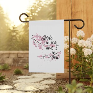 May include: A white garden flag with the text "Abide in me, and I in you." features a floral design of pink cherry blossoms. The flag is displayed on a black metal stand in a garden setting.