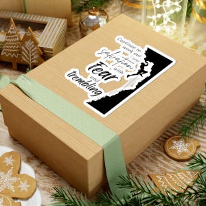 May include: A sticker with the text "Continue to work out your salvation with fear and trembling" and a graphic of a person climbing a mountain. The sticker is on a wrapped gift box with a green ribbon.