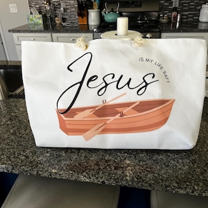 May include: A white canvas tote bag featuring a graphic of a brown rowboat with two oars. The text "Jesus IS MY LIFE RAFT" is printed in black lettering above the boat. The bag includes two thick, knotted rope handles at the top for carrying.