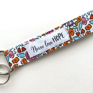 May include: A floral patterned keychain wristlet with a white background. The fabric features orange, pink, and blue flowers. A white label with the words "Never lose HOPE" is sewn onto the wristlet. Two silver key rings are attached.