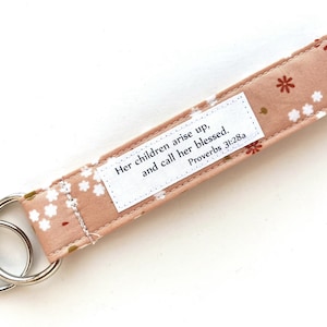 May include: A pink fabric keychain with a white floral pattern and a silver key ring. The keychain has a white label with the text "Her children arise up, and call her blessed. Proverbs 31:28a"