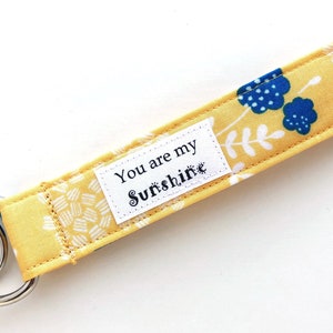 May include: A yellow fabric keychain wristlet with a floral pattern and silver key rings. The wristlet has a white label with the text "You are my Sunshine". The design includes blue flowers and white leaf accents.