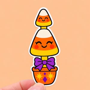May include: A sticker featuring two stacked candy corn shapes with smiling faces, rosy cheeks, and closed eyes. The bottom candy corn sits in an orange pot decorated with yellow starbursts and a purple diamond, accented by a purple bow.