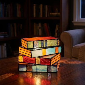 Könnte beinhalten: Ein Stapel von vier Lampen in Buchform aus Buntglas auf einem Holztisch. Die Lampen weisen leuchtende Farben wie Orange, Grün, Blau und Rot auf. Der Hintergrund umfasst ein Bücherregal, ein Fenster und einen gemütlichen Sessel.
