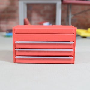 Puede incluir: Un cofre de herramientas rojo en miniatura con cuatro cajones y un compartimento superior. Los cajones tienen detalles blancos. El cofre de herramientas está sobre una superficie blanca.