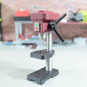 May include: A miniature burgundy drill press with a black handle and base. The drill press has a silver column and a black work surface. The background includes a brick wall and other workshop tools.