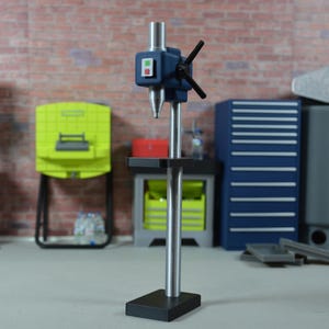 May include: A model workshop with a blue drill press as the focal point, alongside a grey bandsaw, a yellow cabinet, and a blue tool chest. The drill press has a black handle and red and green buttons. The backdrop is a brick wall.