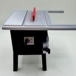 May include: A miniature table saw model featuring a silver top surface with a red blade, a black base, and a control panel on the front. The control panel includes a black dial, a green square button, and a red square button, all mounted on legs.