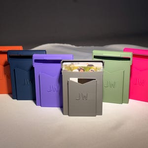 May include: A collection of six rectangular card holders in orange, navy blue, purple, gray, light green, and bright pink. Each holder features a front pocket with the embossed letters JW and a top slot containing visible white cards.