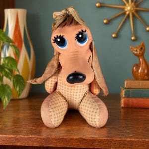 May include: A plush dog toy with a tan and white checkered body, long floppy ears, and a smooth tan face. The dog features large blue eyes with black eyelashes, a black nose, and a tan fabric bow tied at the top of its head. The toy is seated.